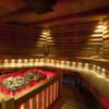 Wood-paneled sauna interior with multiple benches and a red-glowing heater stones on a metal rack in the corner, wooden bucket and ladle visible nearby.