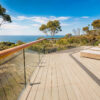 Coastal deck with wicker lounge chairs overlooking the ocean, glass railing, and trees under a blue sky.
