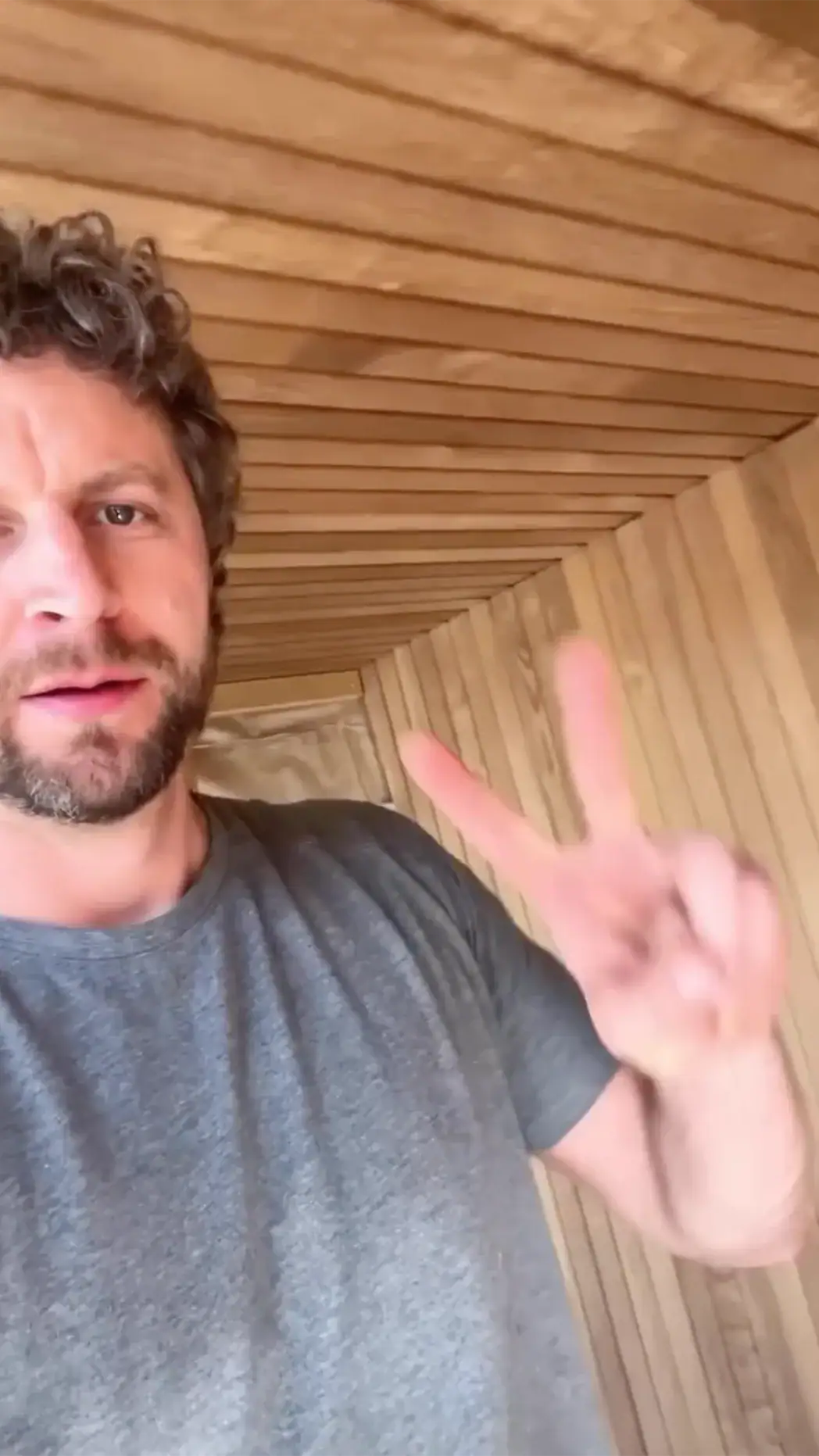 Man with dark hair and beard takes a selfie in a wood-paneled hallway, raising two fingers in a peace sign.