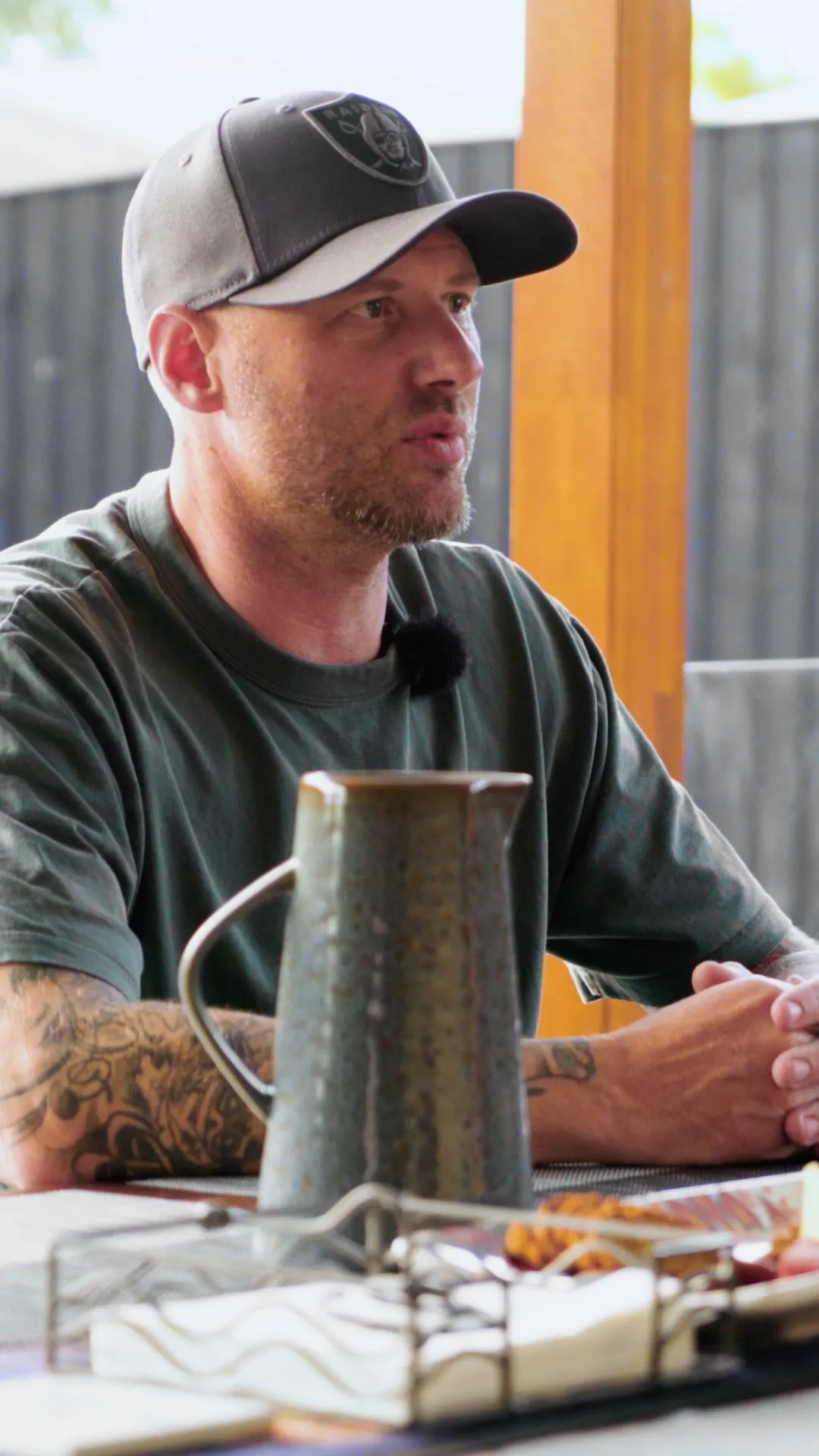 Man in a dark baseball cap and green shirt speaks during a discussion at a table, a mug and objects in foreground.
