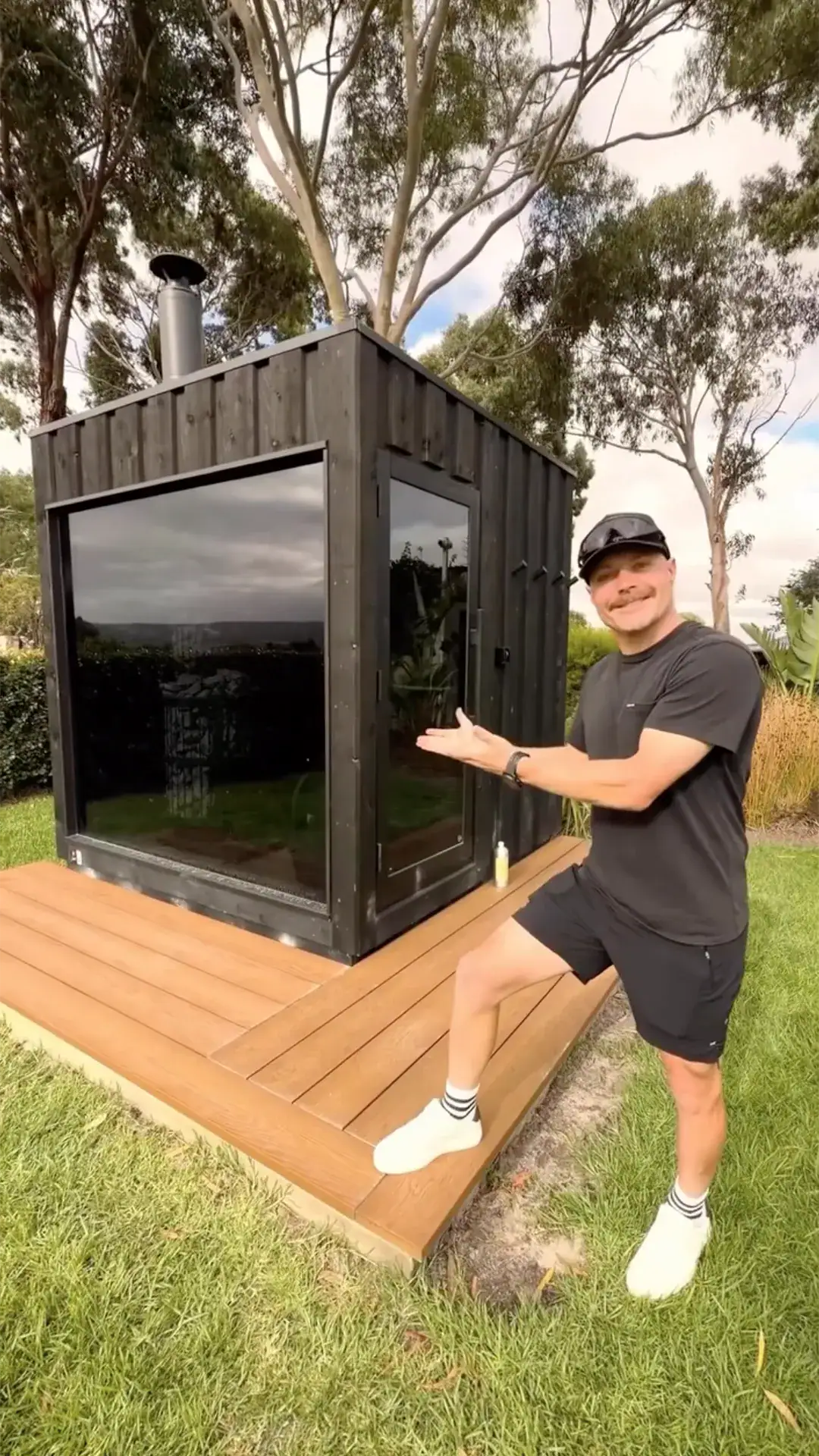 Smiling man in a black shirt, cap, and white sneakers presenting a black wooden tiny house with glass doors on a wooden deck in a backyard.
