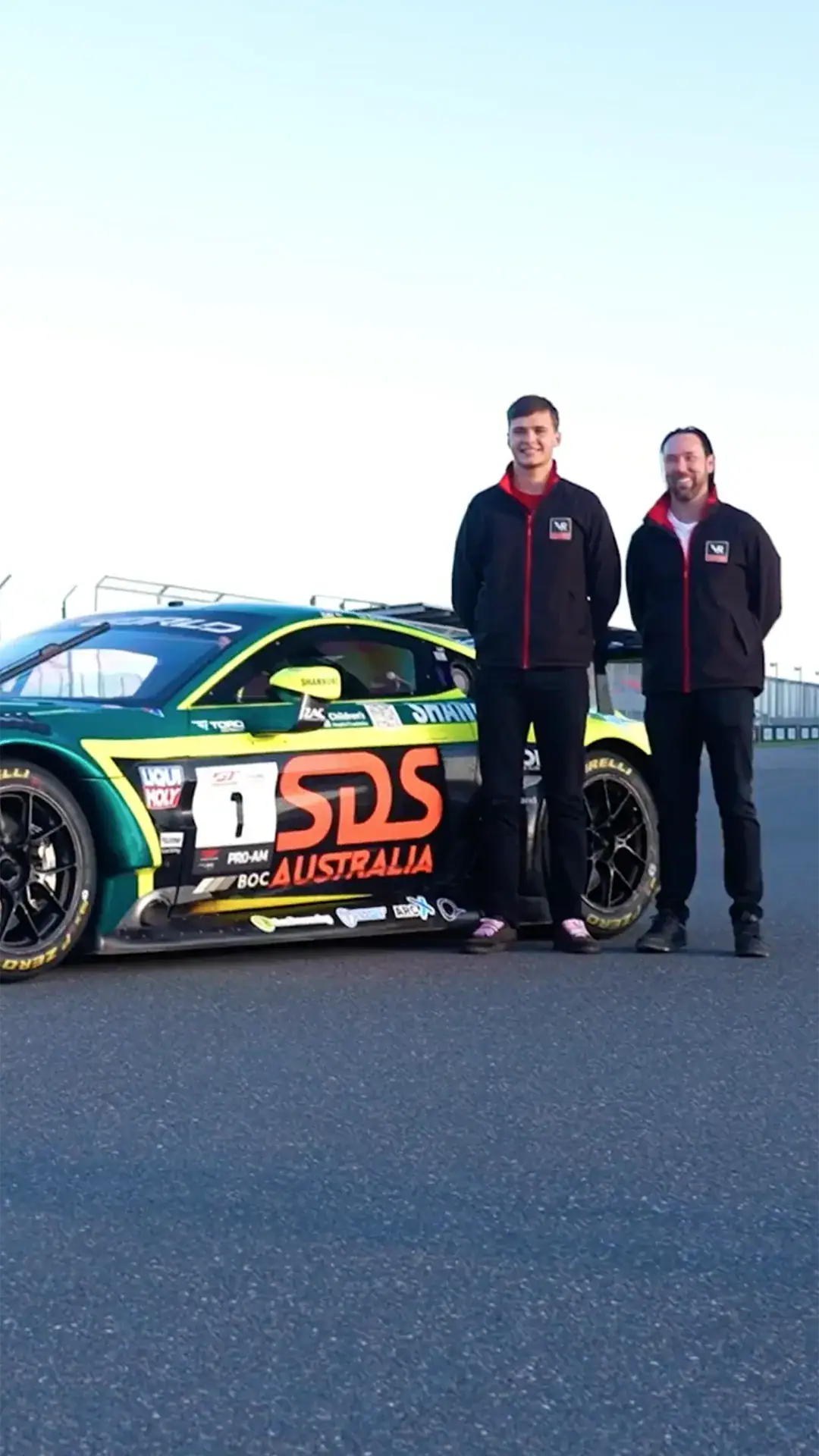 Two men in black jackets with red zippers stand beside a colorful SDS Australia race car on a track surface.