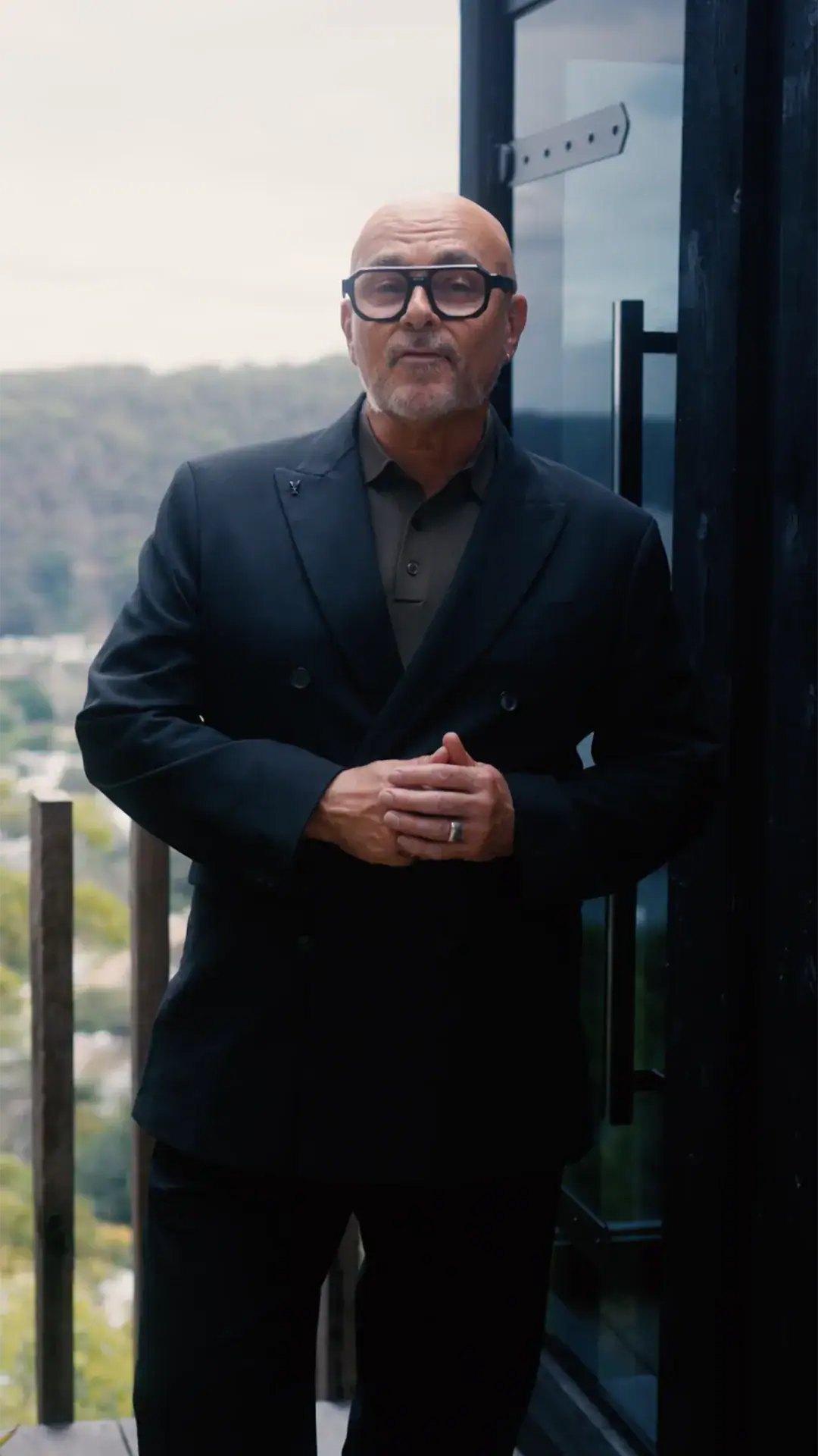 Portrait of a bald man with glasses in a dark suit standing on a balcony, hands clasped in front of him.