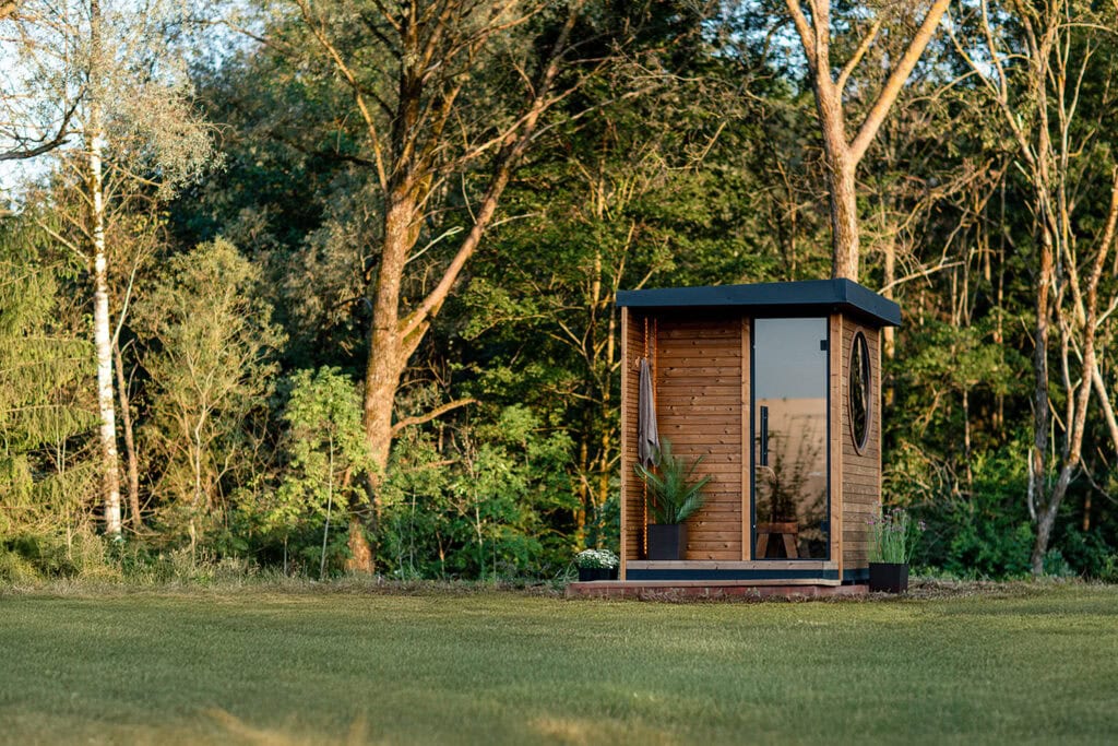 Small wooden cabin with a glass door and circular window in a grassy garden with trees nearby