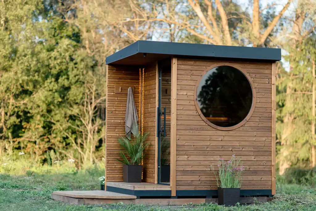 Small wooden cabin with a large circular window, standing on a raised deck in a grassy yard; plants in pots nearby.