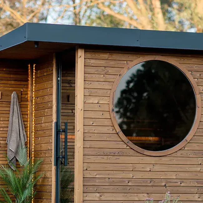 Small wooden cabin with a large circular window, standing on a raised deck in a grassy yard; plants in pots nearby.